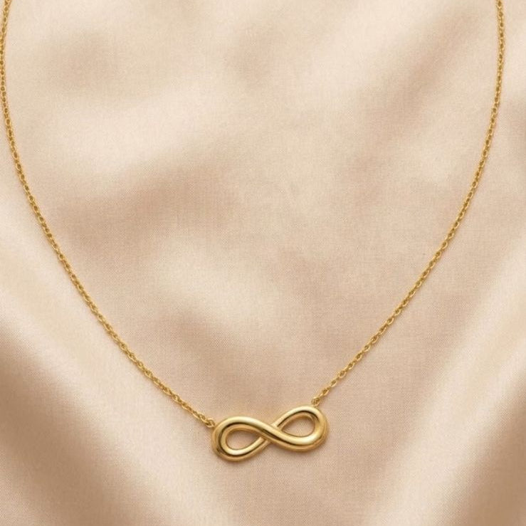 A delicate gold chain necklace featuring a small infinity symbol pendant, displayed on a soft beige satin fabric background.