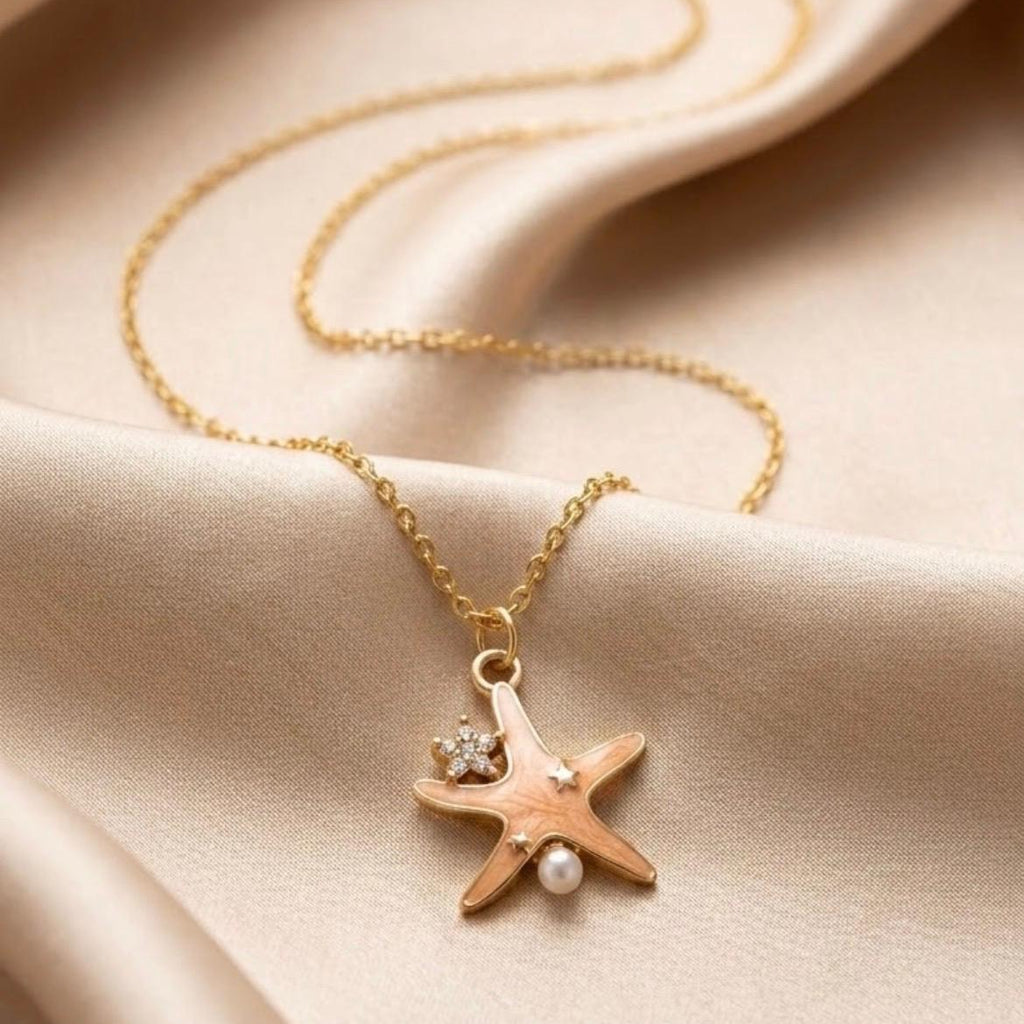 A gold chain necklace with a starfish pendant, styled on draped beige satin fabric. The starfish pendant features a small clear rhinestone and a white pearl.