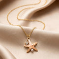 A gold chain necklace with a starfish pendant, styled on draped beige satin fabric. The starfish pendant features a small clear rhinestone and a white pearl.