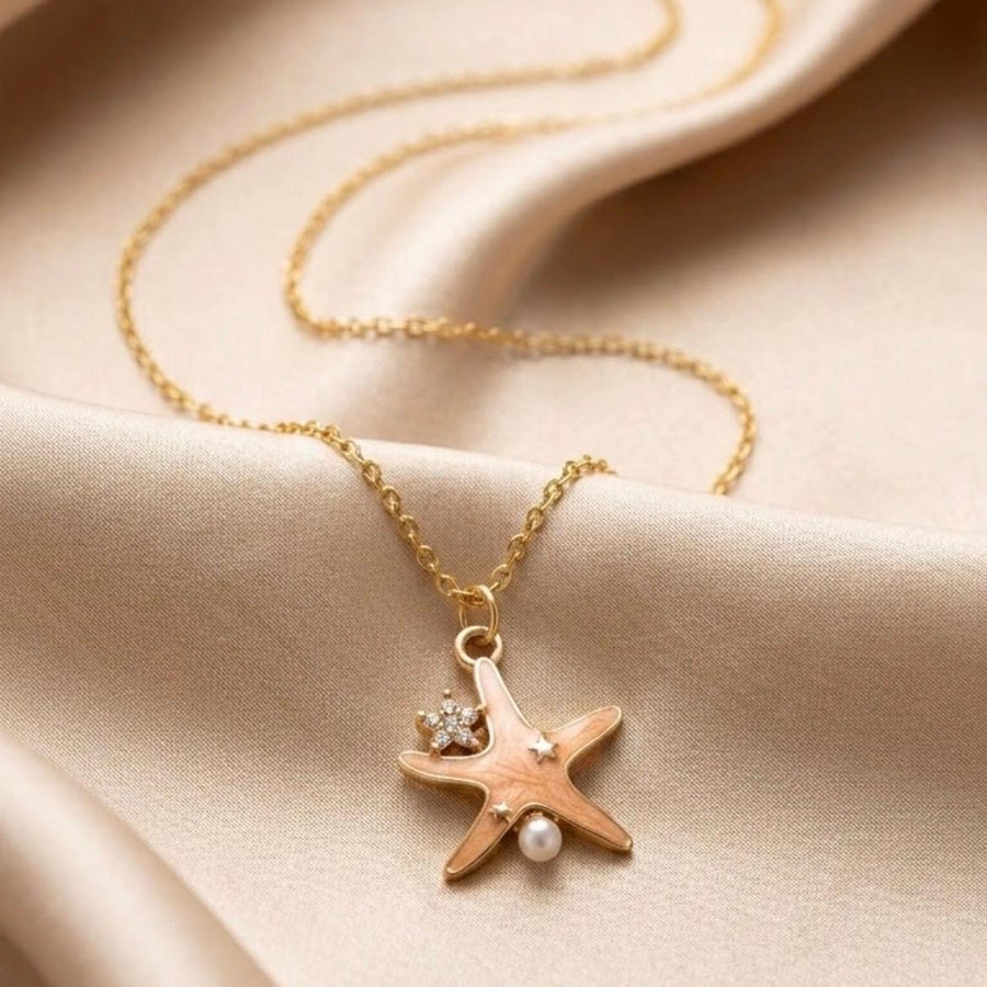 A gold chain necklace with a starfish pendant, styled on draped beige satin fabric. The starfish pendant features a small clear rhinestone and a white pearl.
