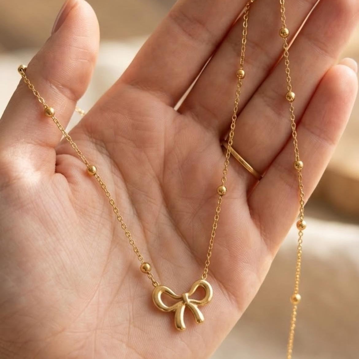 A delicate gold necklace with a small bow pendant and tiny bead accents, held in a person's hand against a soft, warm, blurred background.