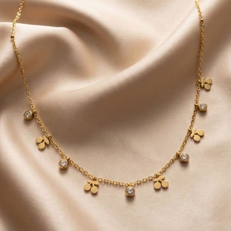 A delicate gold chain necklace with small clover-shaped charms, some adorned with clear stones, displayed on draped beige satin fabric.